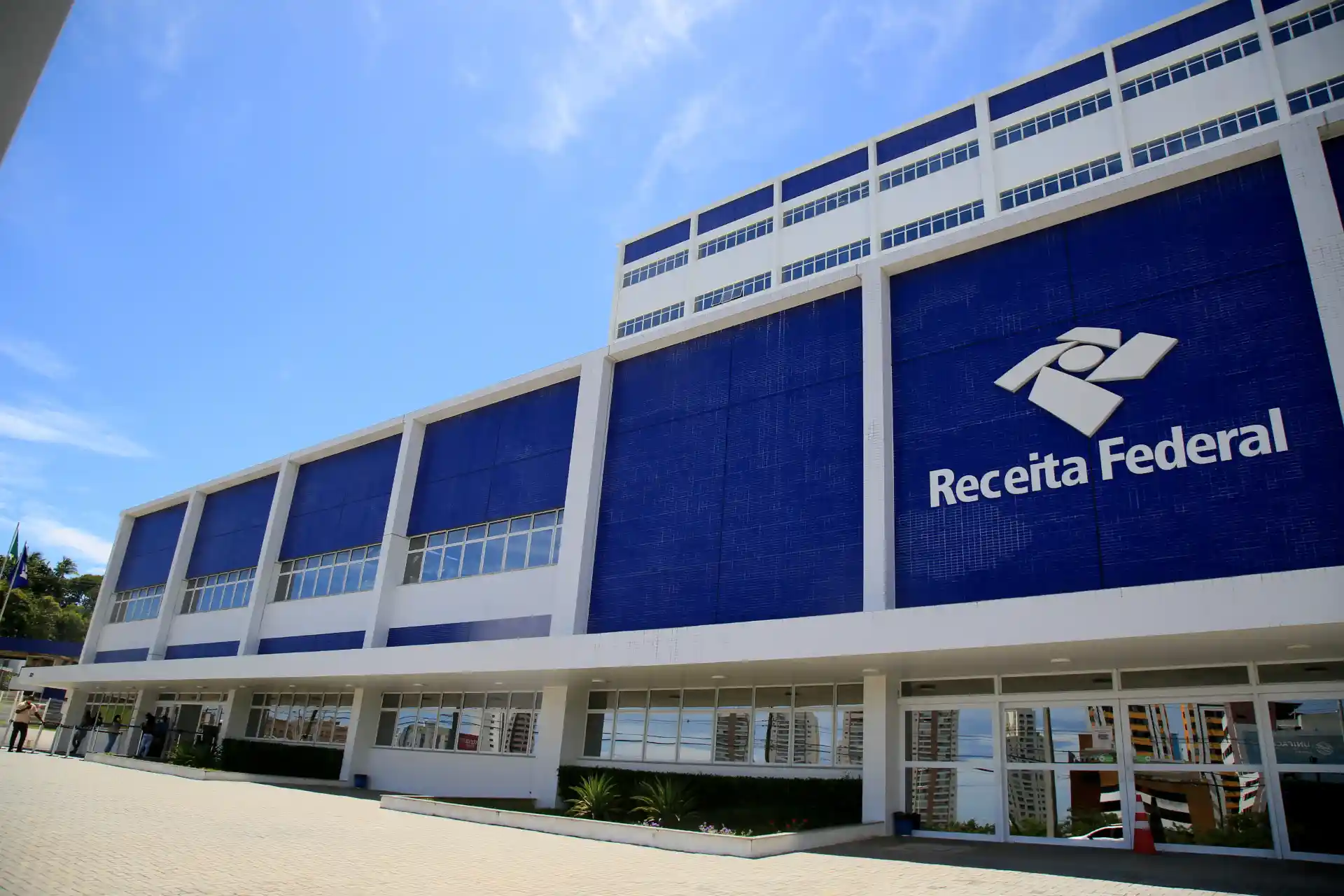 Imagem da fachada da Receita Federal, destacando a entrada principal e o logo da instituição. O céu azul completa a cena, indicando um dia ensolarado.